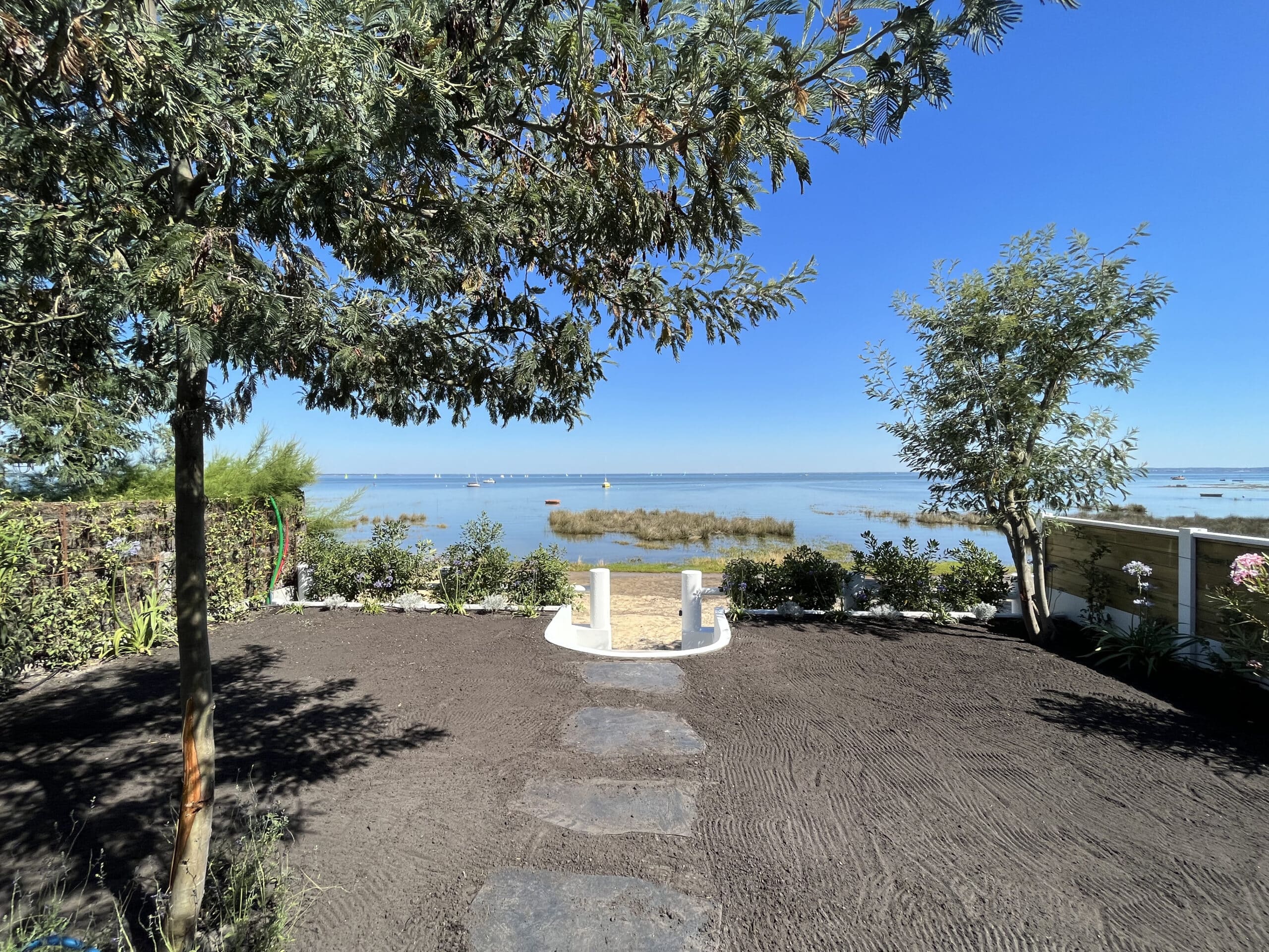 Aménagement de jardin en bord de mer. Terre labourée et pas japonais mènent à une vaste lagune bleue parsemée de voiliers sous un ciel clair.