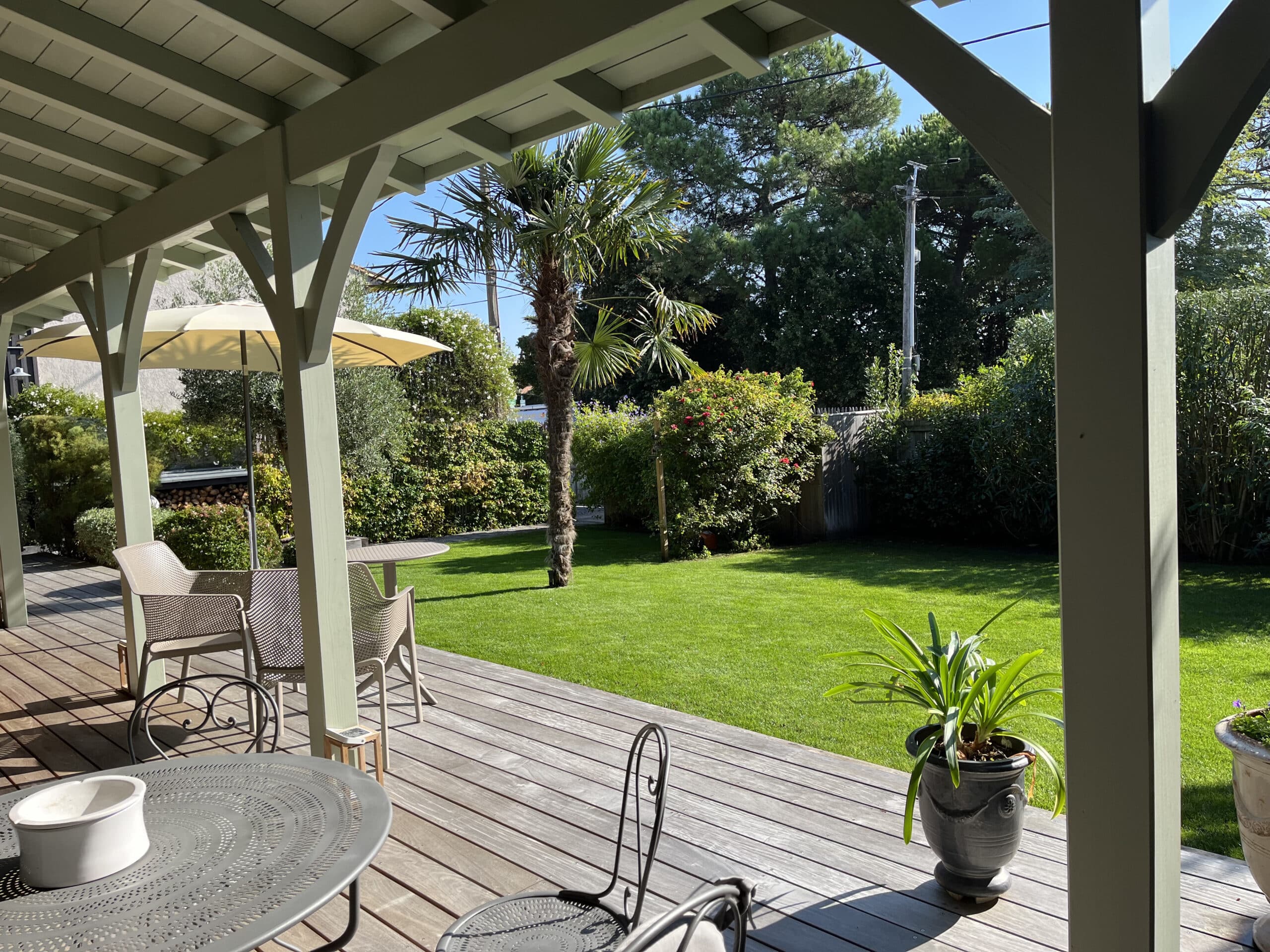 Pergola sur terrasse en bois avec mobilier extérieur, dominant une pelouse verte et un jardin. Palmier, haies et ciel bleu ensoleillé.