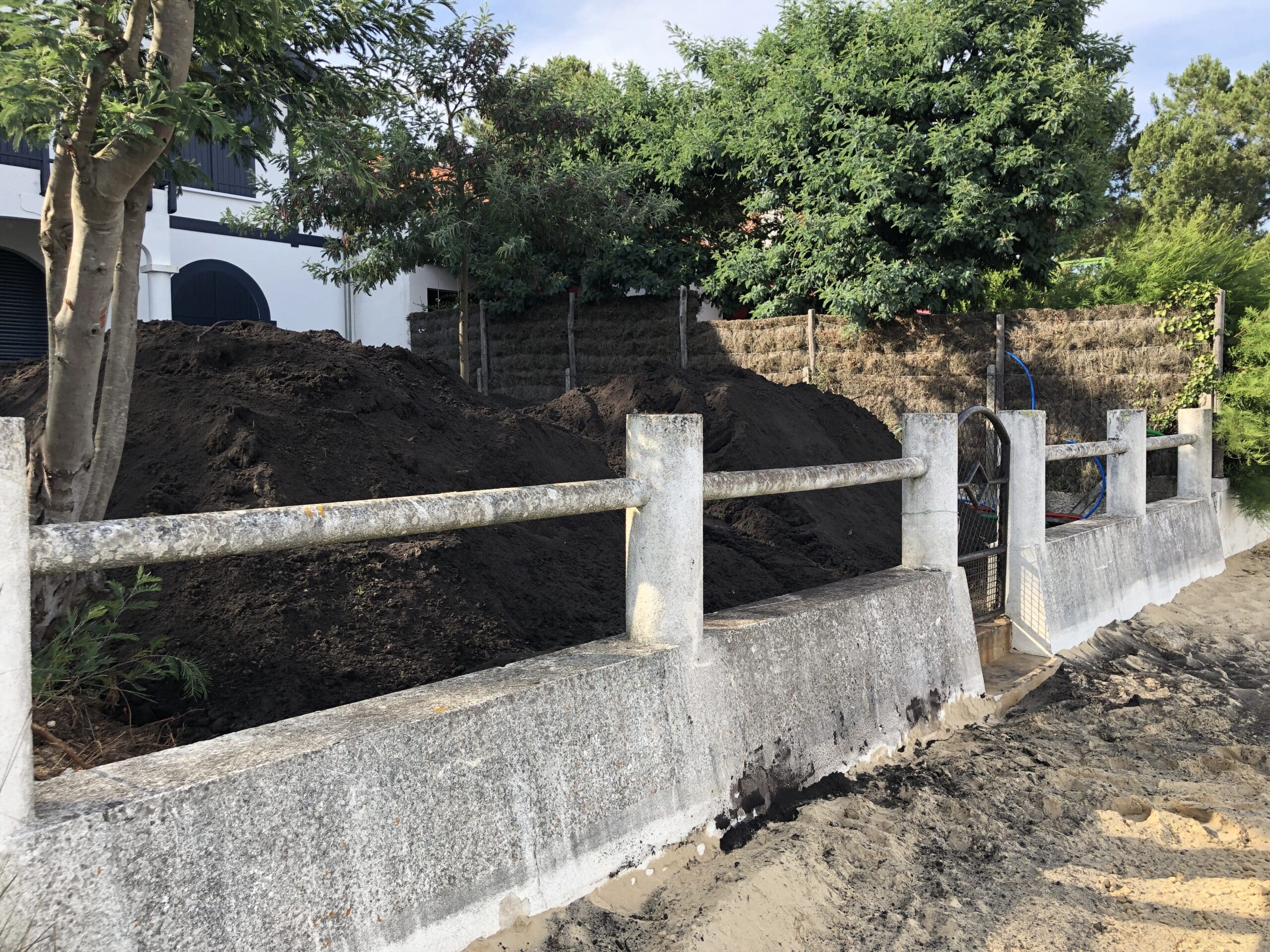 Grand tas de terre végétale noire devant une villa blanche et des arbres. Clôture basse en béton rugueux près d'un sol sablonneux.